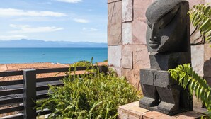 Eine Steinskulptur eines Gesichtes, eine hölzerne Bank und der Blick auf Meer und Berge.