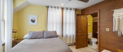 Een slaapkamer met een bed, een plafondventilator, een raam met gordijnen, een nachtkastje met een lamp en een deur die leidt naar een badkamer.