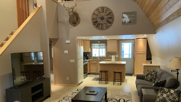A spacious living area with a large TV, a kitchen with wooden cabinets, a dining area with stools, and a staircase leading to an upper level.