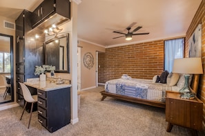 A bedroom with a bed, a desk with a mirror, a ceiling fan, and a brick wall.