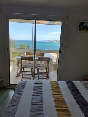 A bedroom with a bed, a view of the sea through a sliding door, and a table with chairs outside.