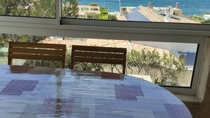 Una mesa redonda con mantel estampado, dos sillas de madera y vista al mar y edificios a través de una ventana.
