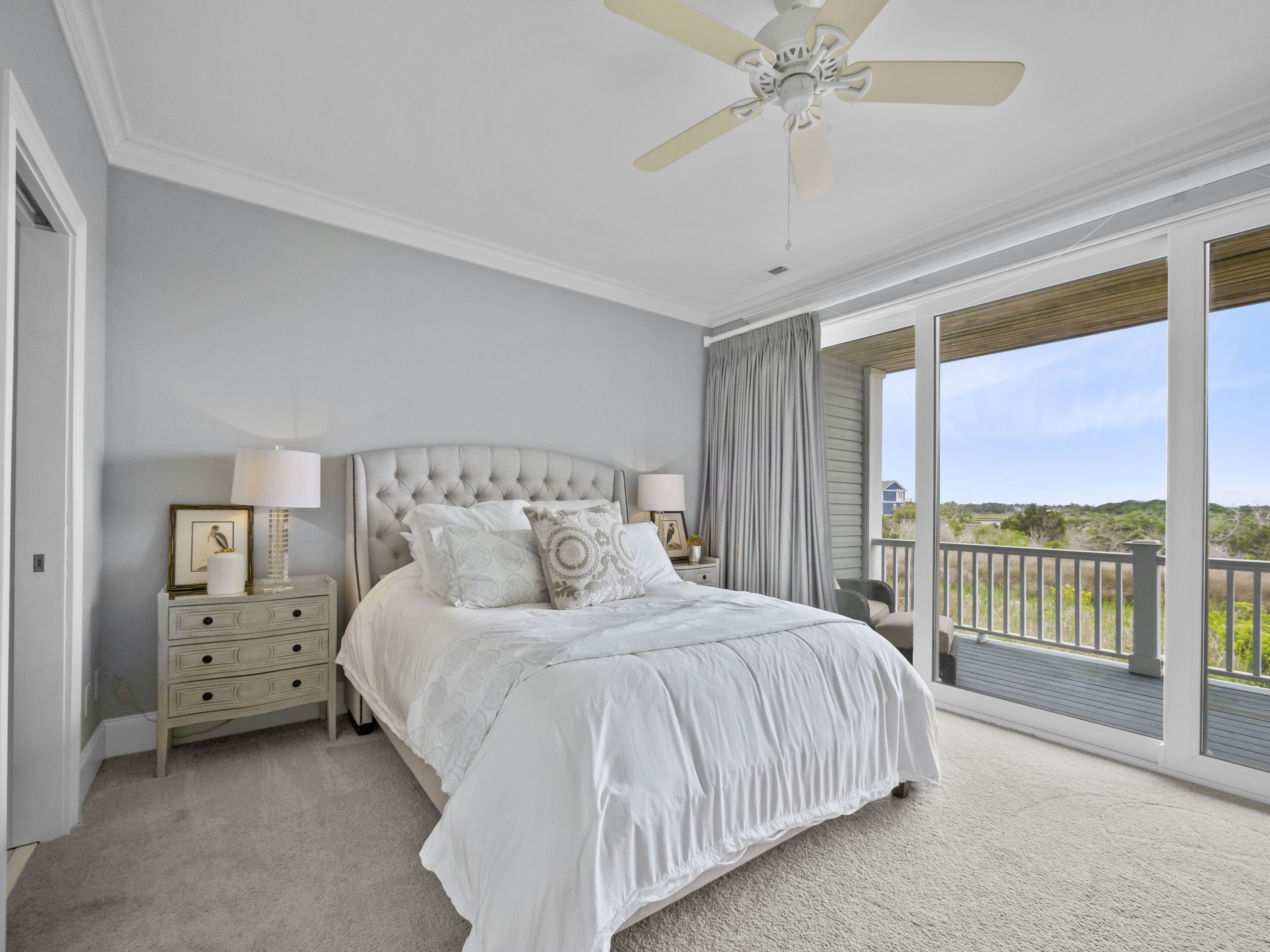 A bedroom with a large bed, a ceiling fan, a nightstand with lamps, and a view of the outdoors through a sliding glass door.