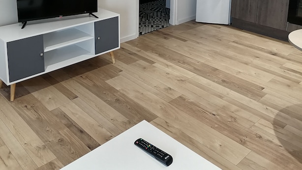 Un salon moderne avec un téléviseur à écran plat posé sur une console blanche, une télécommande sur une table blanche, et un coin cuisine doté de meubles en bois et d’un réfrigérateur blanc.