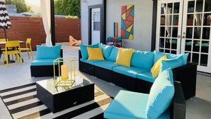 A covered outdoor seating area with a blue sofa, yellow chairs, and a striped rug.