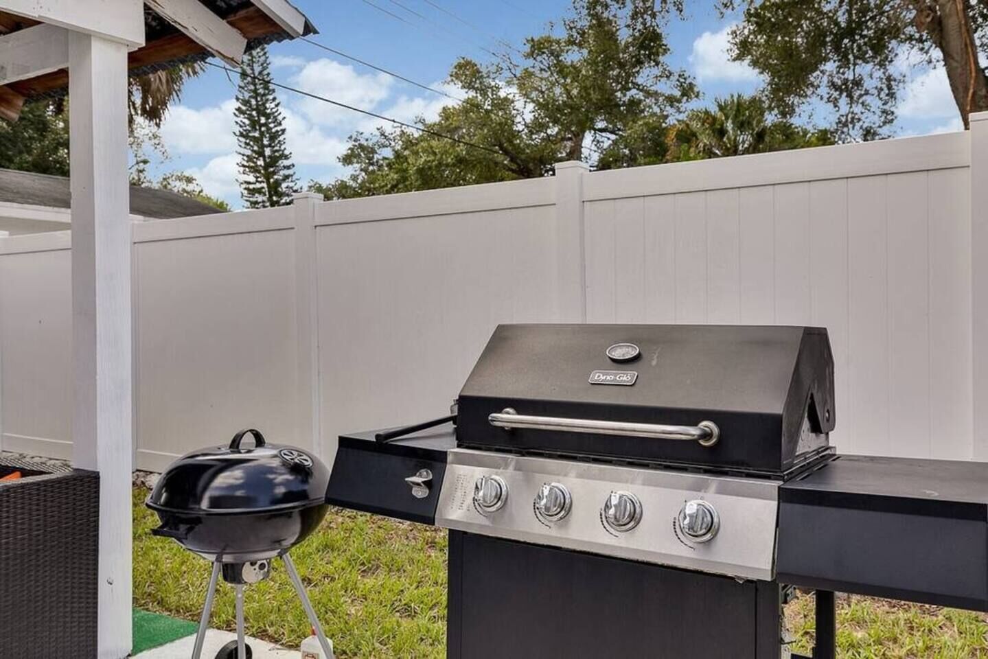 A portable grill with four burners and a side burner, placed outdoors with a white fence and trees in the background.