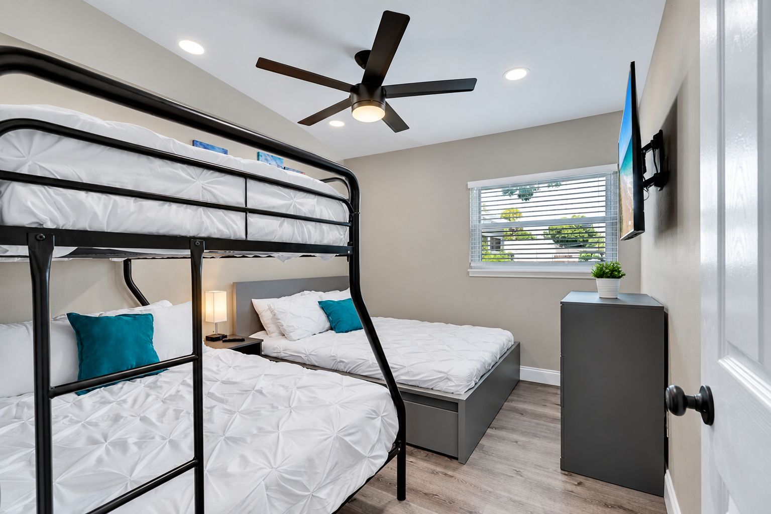 A bunk bed room with a ceiling fan, a bedside table with a lamp, and a window with blinds.