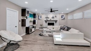 A modern living room with a white sofa, a white armchair, and a coffee table. There is a fireplace with decorative items on the mantel. - Heated Pool & Game Room, Sleeps 14 | 10 Min to Tampa Airport & Stadium (Tampa)