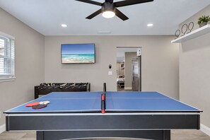 A room with a ping pong table, a foosball table, a ceiling fan, and a wall-mounted TV displaying a beach scene. - Heated Pool & Game Room, Sleeps 14 | 10 Min to Tampa Airport & Stadium (Tampa)