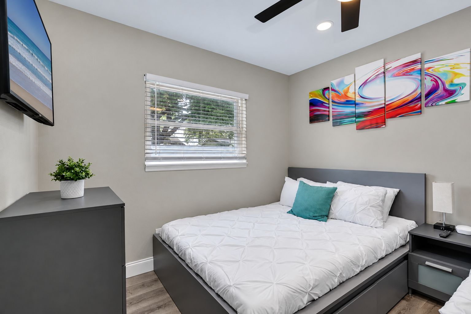 A modern bedroom with a bed, a nightstand, a lamp, a TV, a window with blinds, and a wall with abstract art.