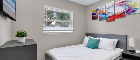 A modern bedroom with a bed, a nightstand, a lamp, a TV, a window with blinds, and a wall with abstract art.
