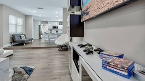 A modern living room with a flat-screen TV mounted on the wall, a gaming chair, and a console on a white table. - Heated Pool & Game Room, Sleeps 14 | 10 Min to Tampa Airport & Stadium (Tampa)