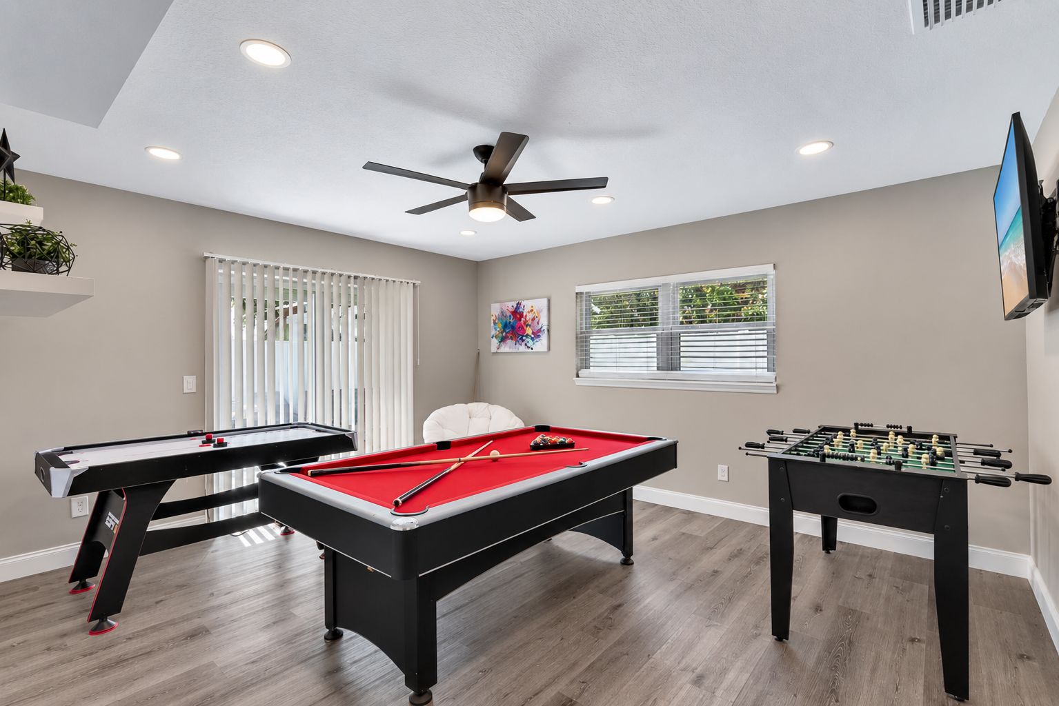 Une pièce équipée d’une table de billard, d’un baby-foot, d’un ventilateur de plafond et d’une fenêtre avec des stores.