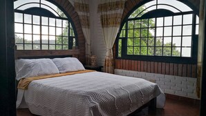 A bed with a white quilt and pillows, a wooden headboard, and a view through arched windows to greenery outside.