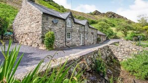 A stone house with a sloped roof, a stone pathway leading to the entrance, and a hillside backdrop.