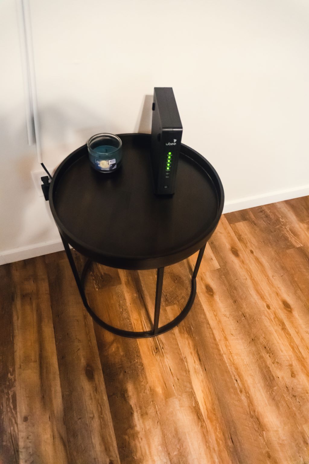 Een klein, rond tafeltje met een zwart metalen frame, een zwart elektronisch apparaat met groene lampjes en een klein glazen recipiënt op tafel.