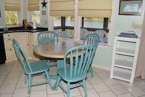A dining area with a round table and four turquoise chairs, a white cabinet, and a view of a body of water through a window with blinds.
