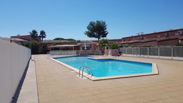 A rectangular swimming pool with a tiled deck, surrounded by a white fence, and adjacent to a building with a tiled roof. - Villa in secure residence with swimming pool 200m from the beach (Saint-Cyprien)