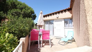 An outdoor patio with two pink chairs and a blue chair, a beige umbrella, and a building with a tiled roof and a white door. - Villa in secure residence with swimming pool 200m from the beach (Saint-Cyprien)
