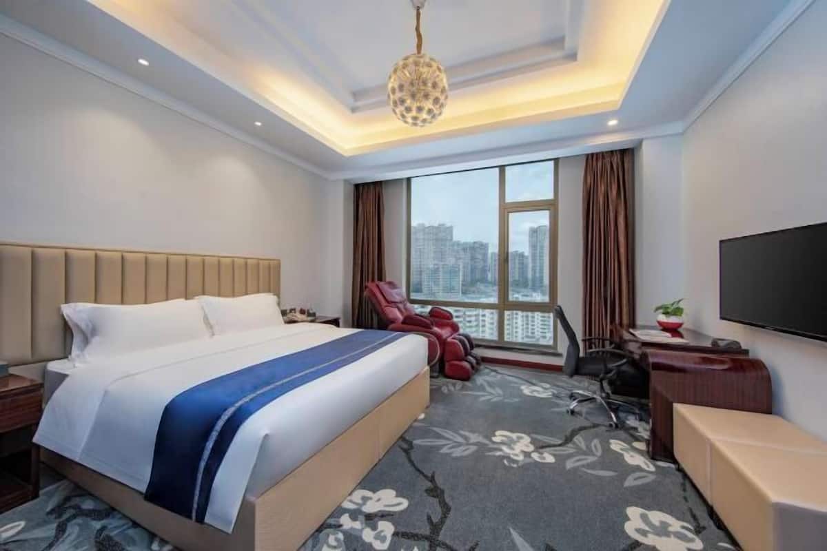 Habitación de hotel con cama, escritorio, silla, sillón de masaje, televisor y una ventana grande con vistas a la ciudad.
