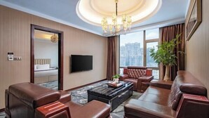 Habitación de hotel con sofá, sillones, mesa de centro, televisor, ventana grande con vista a la ciudad y candelabro.