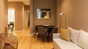 A modern living room with a sofa, a dining table with chairs, a wicker chair, and a painting on the wall.