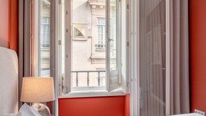 Ein Hotelzimmer mit Bett, einem Fenster mit Blick auf ein Gebäude und einer Lampe auf dem Nachttisch.