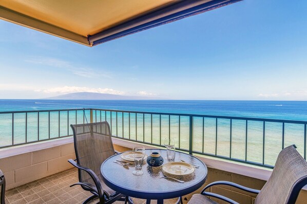 A balcony with a table set for two, overlooking a clear blue sea and a distant mountain range.