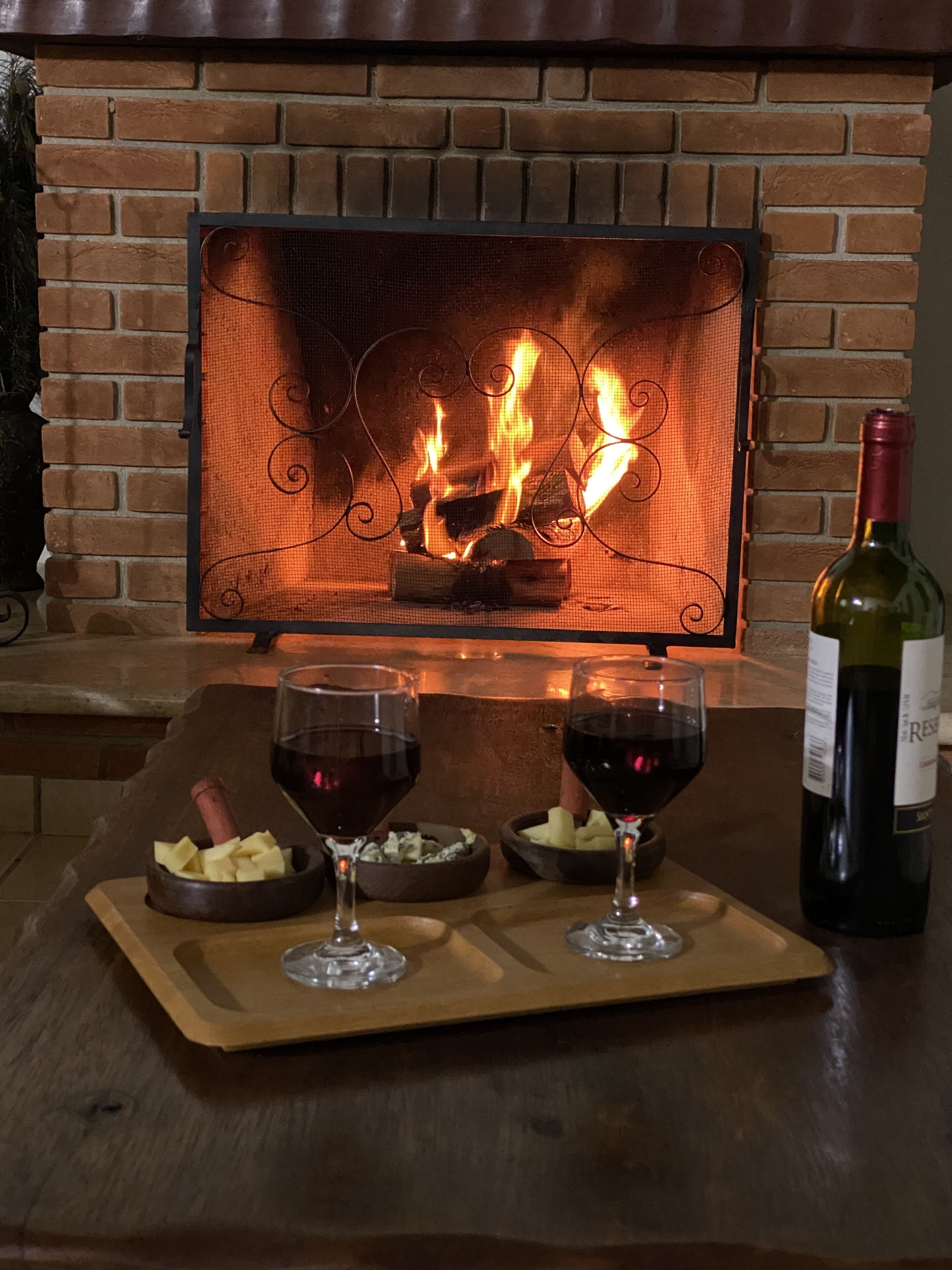 Una chimenea con pared de ladrillo, dos copas de vino tinto, una botella de vino y una bandeja de madera con queso y galletas.