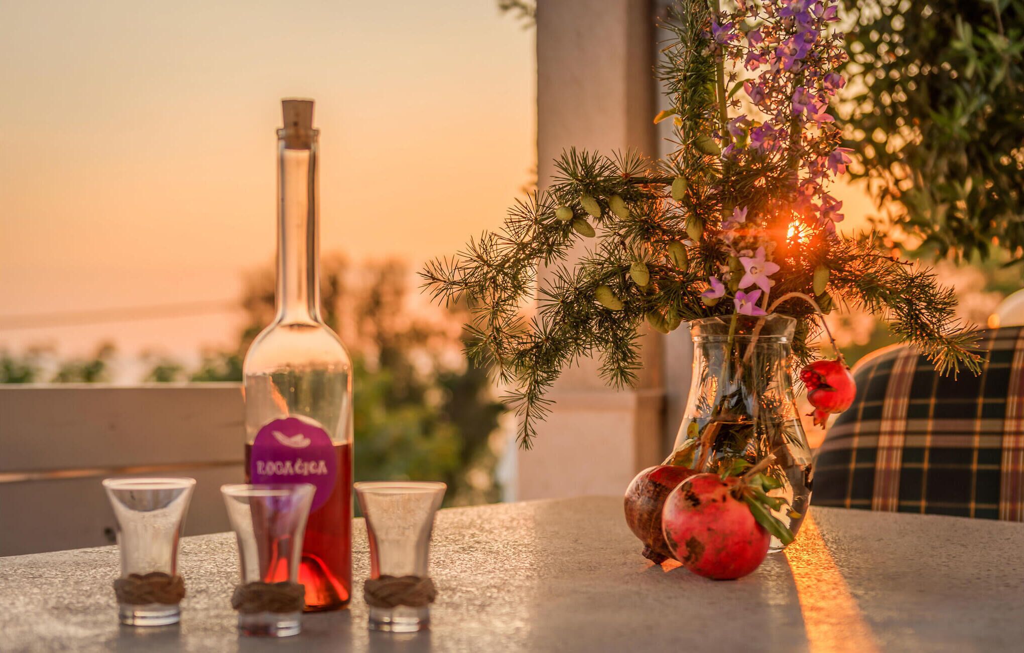 A table with a bottle, shot glasses, and a vase with flowers and a pomegranate.