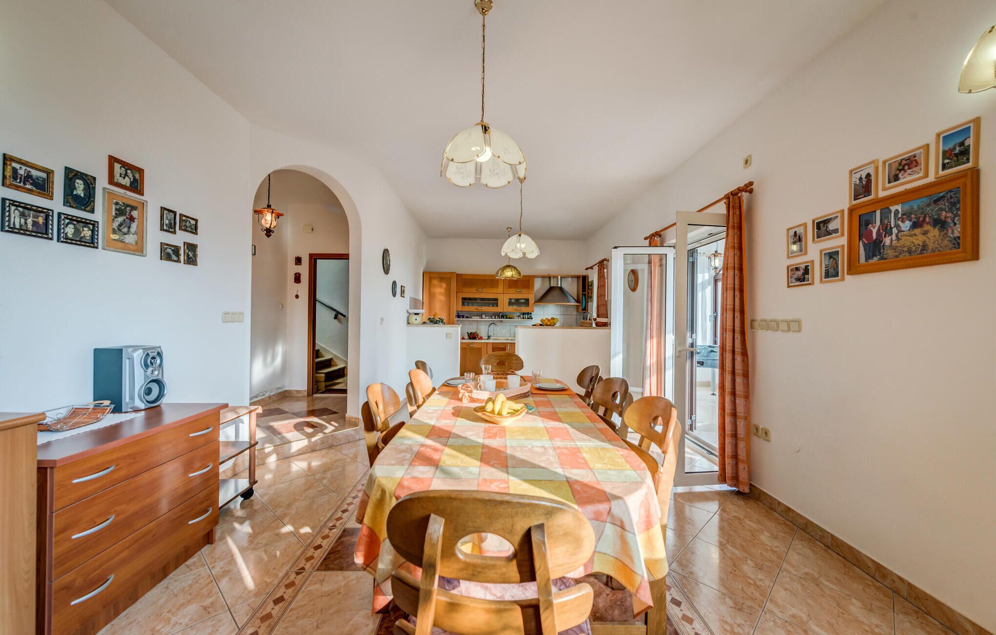 Un'ampia sala da pranzo con un lungo tavolo, sedie in legno e una tovaglia colorata. Si notano anche pensili da cucina e una scala che conduce a un altro piano.
