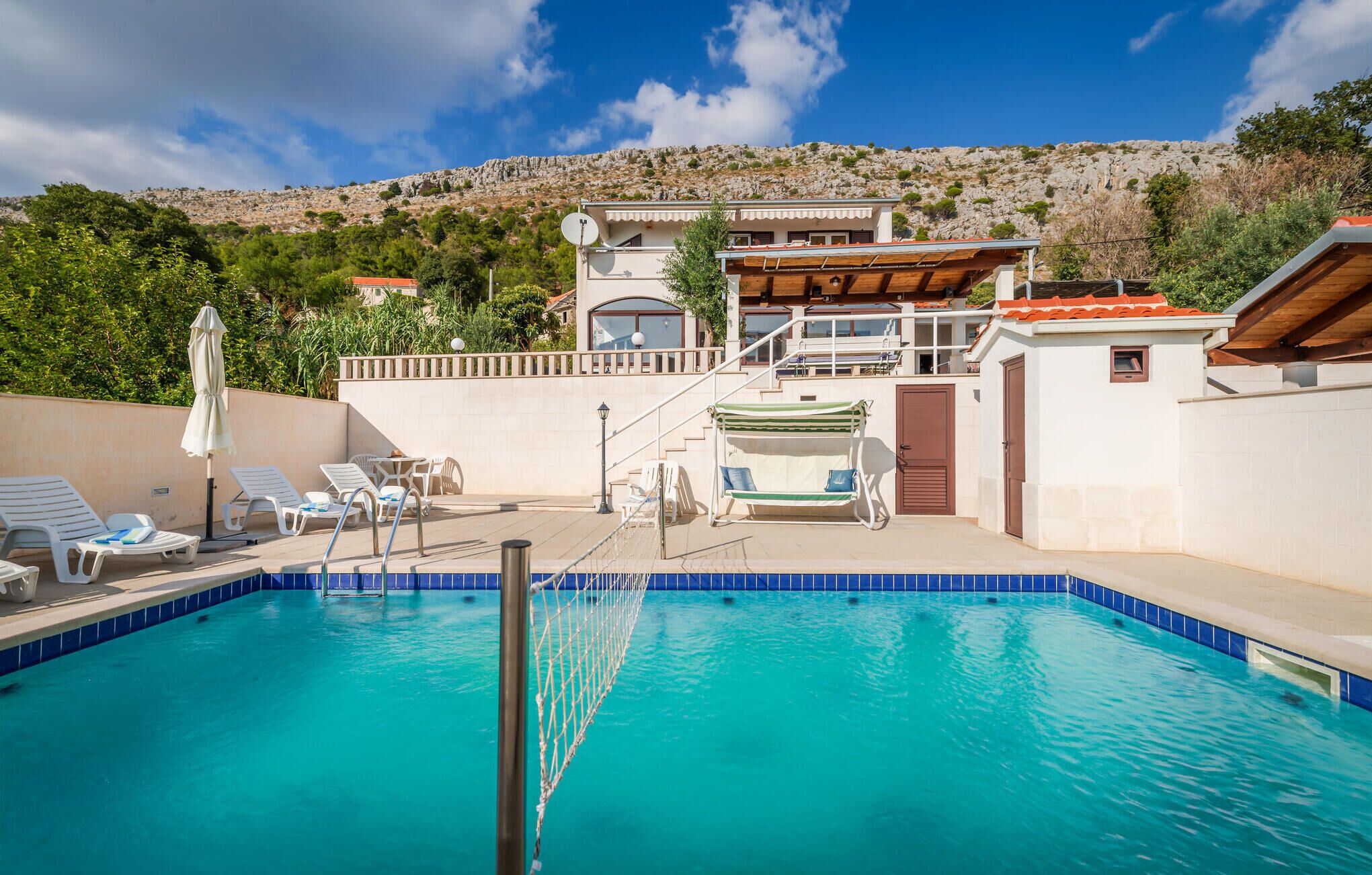Une piscine dotée de chaises longues et d’une zone délimitée par un filet, entourée d’un bâtiment avec un balcon et offrant une vue sur un paysage montagneux.