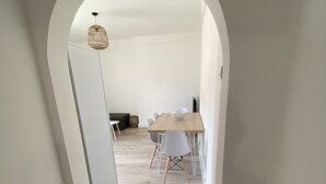 Un couloir mène à une salle à manger où se trouvent une table et des chaises en bois, une porte blanche et un luminaire suspendu.