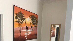 Un couloir orné d’un tableau représentant des girafes et des arbres, avec un miroir et une poubelle.