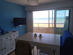A room with a large window offering a beach view, a flat-screen TV mounted on the wall, a white table with a chair, and a blue sofa.