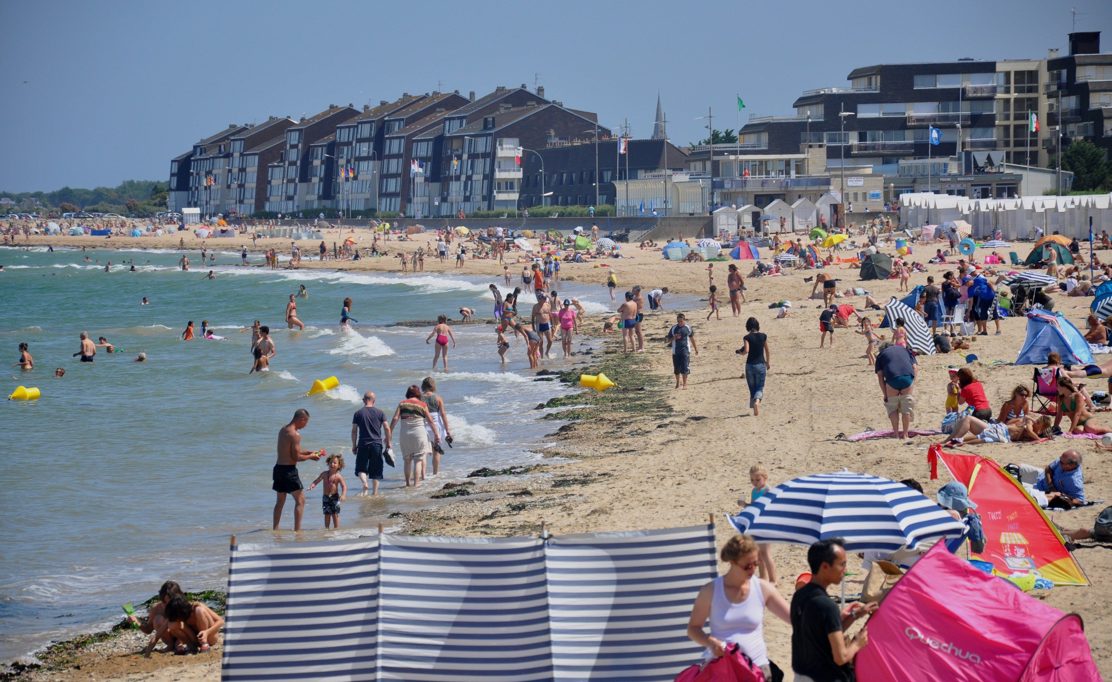 A crowded beach with people enjoying the water and sun. There are buildings in the background and a clear sky.