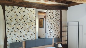 Un dormitorio con techo de madera, una cama con ropa blanca, una mesita de noche con lámpara y una pared con papel tapiz estampado.