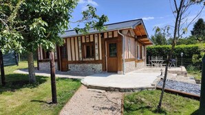 Una cabaña de madera con base de piedra, un sendero de grava que la conduce y un pequeño jardín con árboles y una cerca.