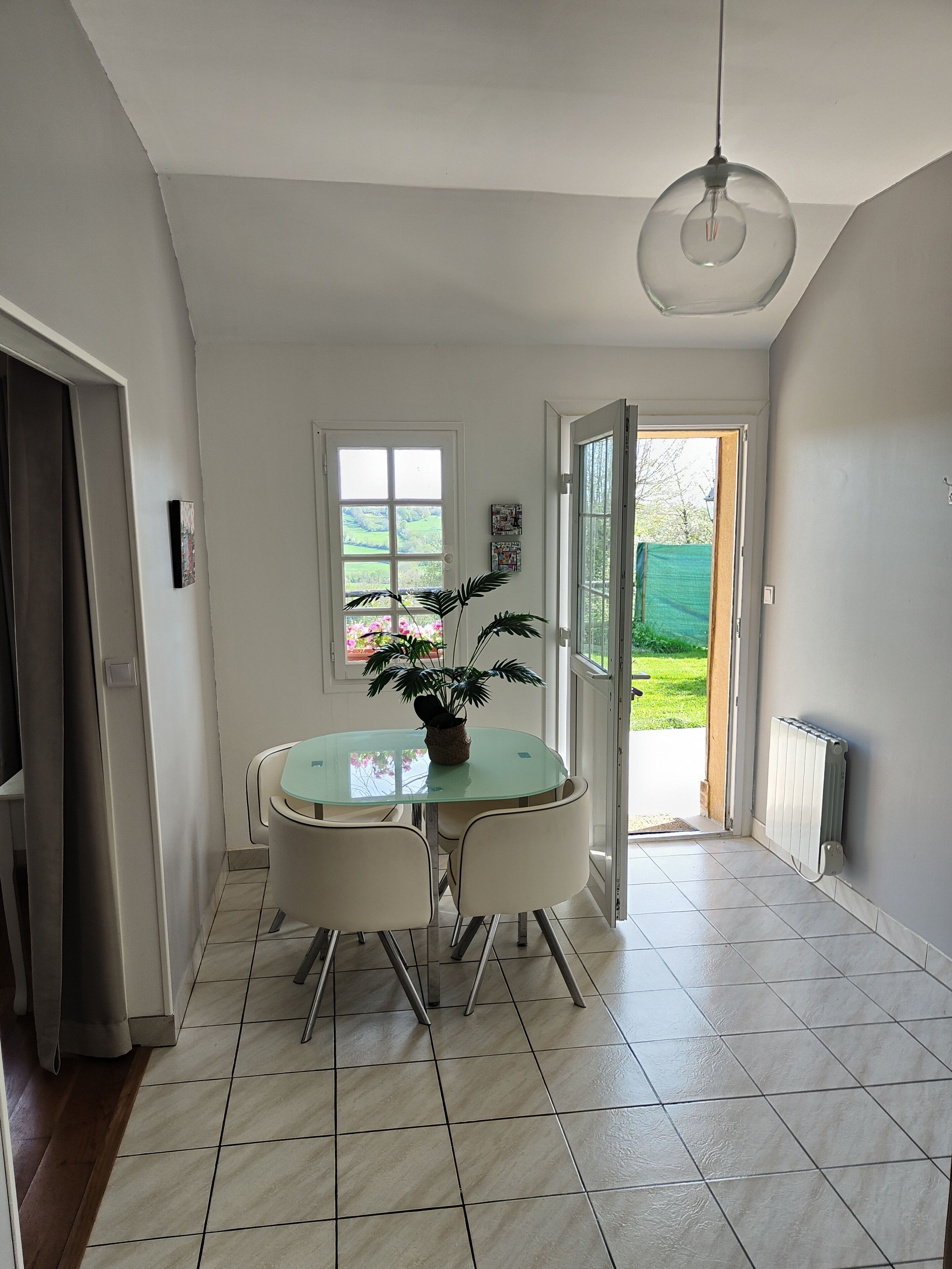 Un coin repas avec une table et des chaises en verre, un luminaire suspendu et une vue sur un jardin par une porte.