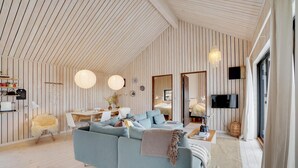 A spacious living room with a high ceiling, wooden panel walls, a large sofa, a dining table, and a TV mounted on the wall.