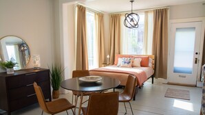 Ein Schlafzimmer mit einem Bett, einem Esstisch mit Stühlen, einem Schminktisch und einem großen Fenster mit Vorhängen.