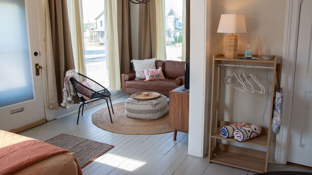 Ein gemütliches Wohnzimmer mit einem braunen Ledersofa, einem runden Hocker, einem Holztisch mit Lampe und einem Regal mit hängenden Gegenständen.