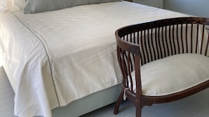 Ein Schlafzimmer mit Bett, Kissen, einem Sessel, einem Nachttisch, einer Lampe und einem Fenster mit Blick ins Grüne.