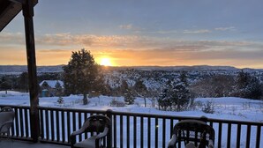 A covered outdoor area with two wicker chairs and a wooden railing, overlooking a snowy landscape with a sunset in the background.