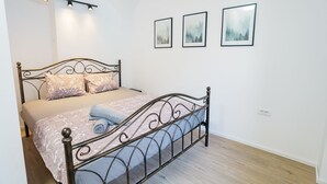 Ein Schlafzimmer mit einem Metallbettgestell, einer beigen Bettdecke und zwei gemusterten Kissen. An der Wand hängen drei gerahmte Bilder und eine Deckenleuchte.
