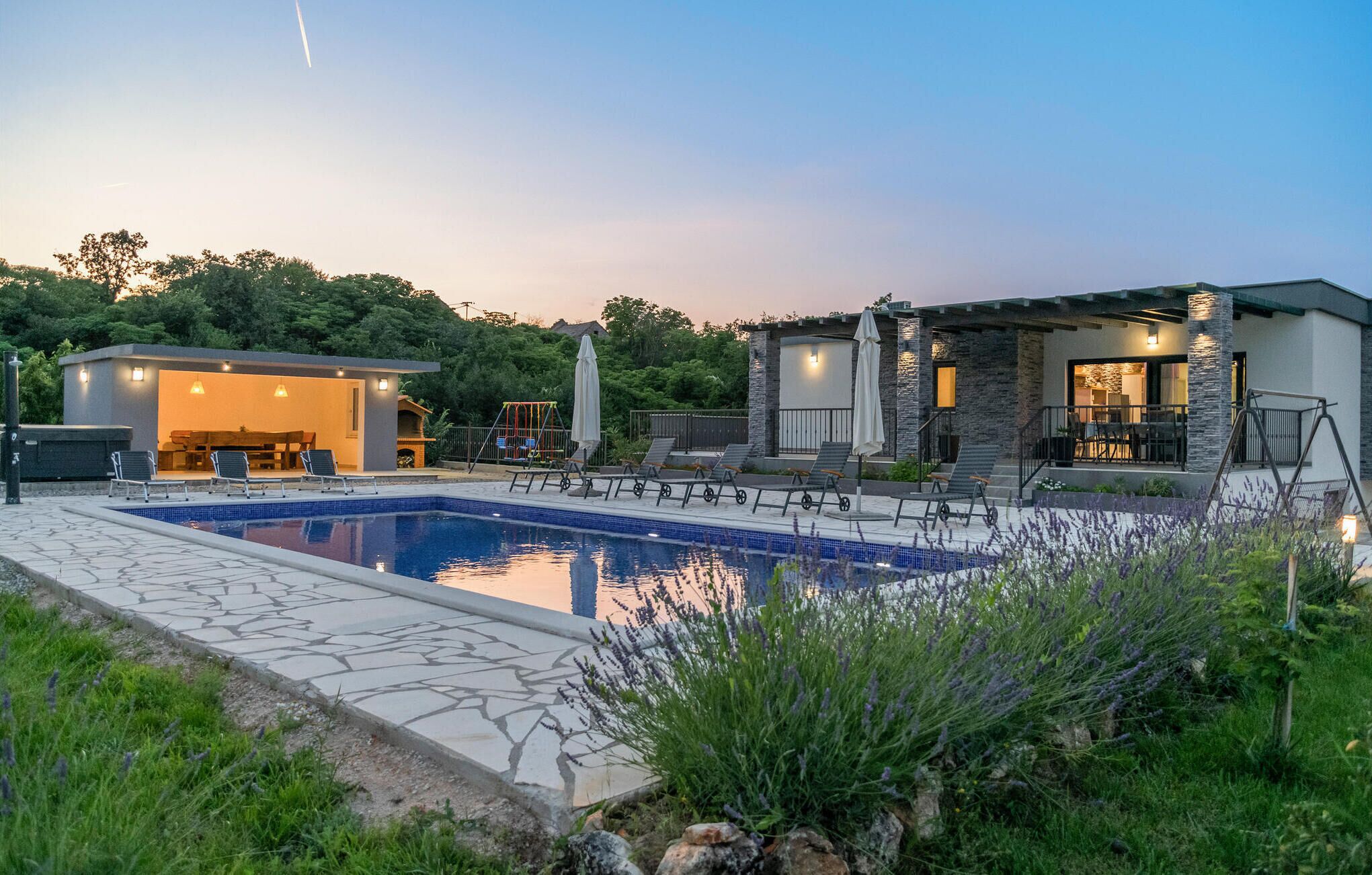 A modern outdoor pool area with a stone deck, lounge chairs, and a small building with a fireplace.