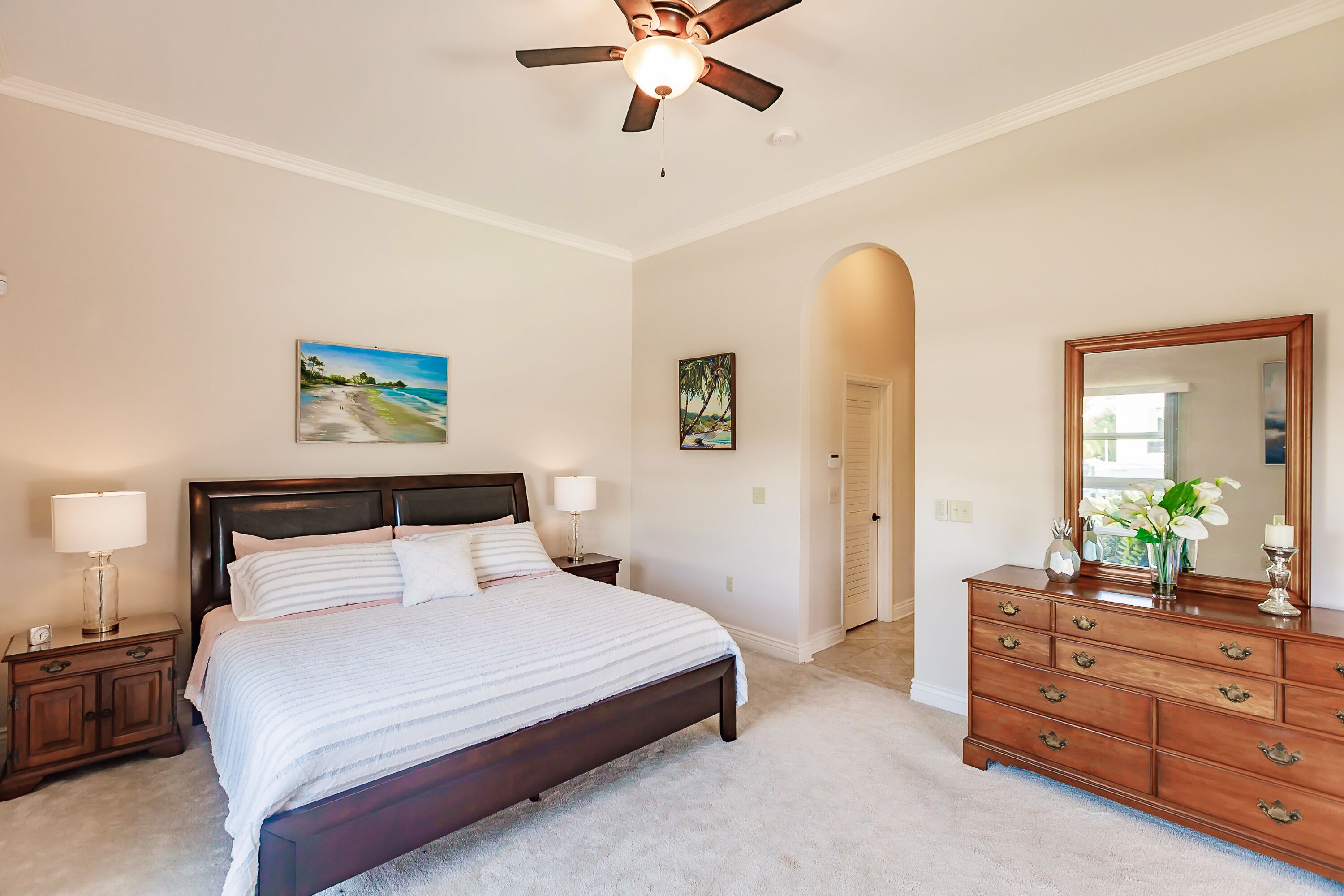 A bedroom with a bed, nightstands, a dresser, a ceiling fan, and two paintings on the wall.