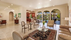 A spacious living room with a large sofa, a coffee table, and a dining area with chairs and a table. There is a kitchen visible through an archway, and a view of a swimming pool through large glass doors. - LUXURY HOUSE BY THE CANAL ☀️ (Siesta Key)
