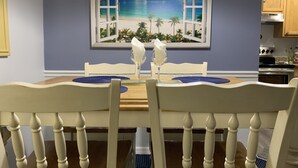 A dining area with a table set for two, a painting of a beach scene, and wooden chairs with a white finish. - Cozy ❤️ Beach Style Condo near Disney & Universal (Kissimmee)