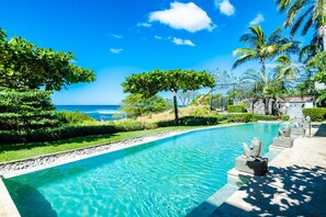A swimming pool with clear blue water, surrounded by greenery and palm trees, with a view of the ocean.